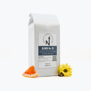 A packaged bag of North Arrow Coffee Company's Blonde No. 20 coffee with a white label, accompanied by a slice of orange and a flower.