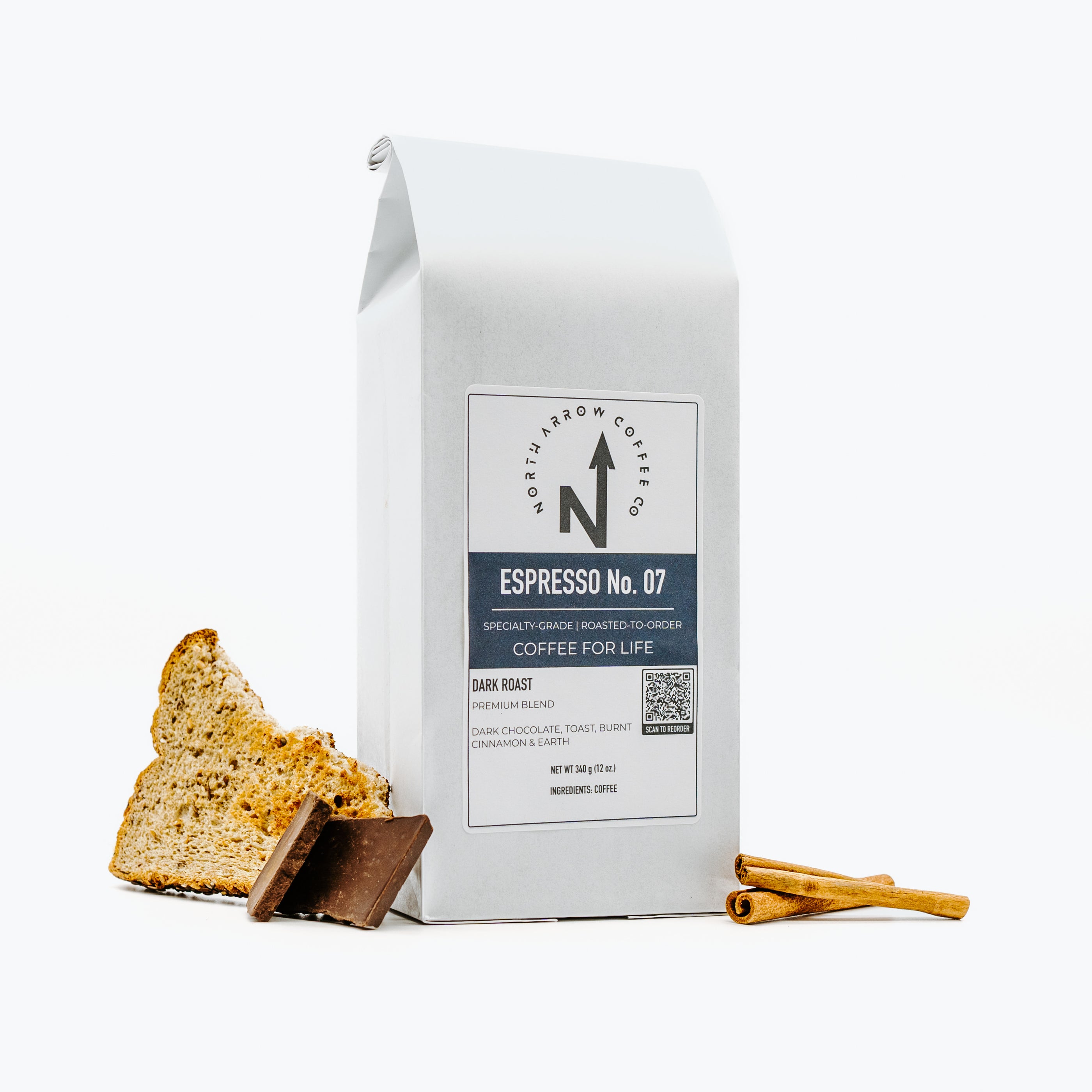 A packaged bag of North Arrow Coffee Company Espresso No. 07 with an indication of being roasted for life, accompanied by a piece of bread and a cinnamon stick, against a white background.